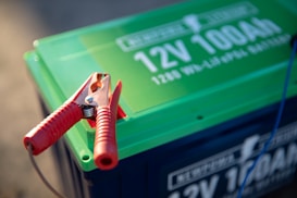 A close-up view of a car battery with a prominent red jumper cable clamp attached to it. The battery has a green top with white text displaying '12V 100Ah'. A cable is seen extending from the clamp, suggesting it is in use.