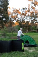 A person is setting up a green tent in an outdoor setting with trees in the background. In the foreground, a portable solar panel is placed on the grass.