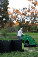 A person is setting up a green tent in an outdoor setting with trees in the background. In the foreground, a portable solar panel is placed on the grass.
