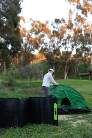 A person is setting up a green tent in an outdoor setting with trees in the background. In the foreground, a portable solar panel is placed on the grass.