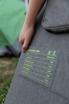 A person holds onto a grey fabric-covered item with printed specifications. The text includes product details such as model and power ratings. The background features a green outdoor setting with grass and part of a tent.