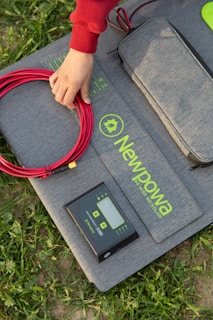 A hand in a red sleeve is arranging red electrical cable on a grey fabric mat. The mat features the branding 'Newpowa' in bright green text. A small electronic device, possibly a solar charge controller, is placed on the mat next to the cable. The scene is set outdoors, as indicated by the grass surrounding the mat.