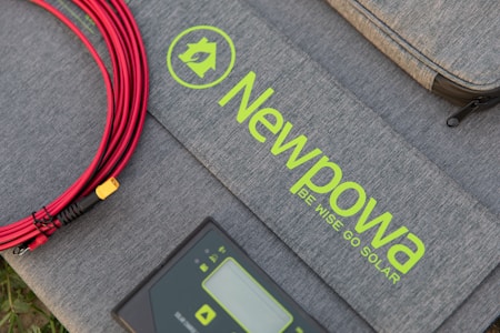 A grey textured surface features a coiled red cable, a grey device with digital display controls, and a soft case with the text 'Newpowa BE WISE GO SOLAR' in bright green lettering.