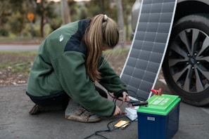 A person crouches on the ground connecting cables from a portable solar panel into a blue and green power pack. The panel is propped up against a car wheel. The setting appears outdoors with trees in the background.