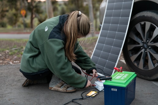 A person crouches on the ground connecting cables from a portable solar panel into a blue and green power pack. The panel is propped up against a car wheel. The setting appears outdoors with trees in the background.