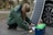 A person crouches on the ground connecting cables from a portable solar panel into a blue and green power pack. The panel is propped up against a car wheel. The setting appears outdoors with trees in the background.