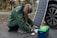A person crouches on the ground connecting cables from a portable solar panel into a blue and green power pack. The panel is propped up against a car wheel. The setting appears outdoors with trees in the background.