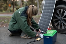 A person crouches on the ground connecting cables from a portable solar panel into a blue and green power pack. The panel is propped up against a car wheel. The setting appears outdoors with trees in the background.