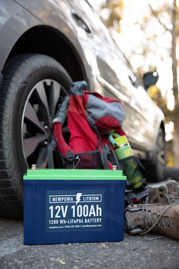 A 12V 100Ah lithium battery is placed on the ground next to a parked car. Nearby, there is a hiking backpack in red and gray, a green water bottle, and a pair of brown hiking boots. The scene suggests a context of outdoor activity or travel.