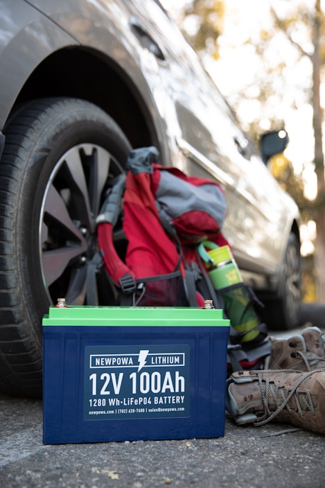 A 12V 100Ah lithium battery is placed on the ground next to a parked car. Nearby, there is a hiking backpack in red and gray, a green water bottle, and a pair of brown hiking boots. The scene suggests a context of outdoor activity or travel.