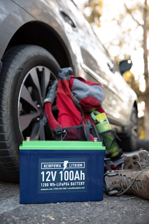 A 12V 100Ah lithium battery is placed on the ground next to a parked car. Nearby, there is a hiking backpack in red and gray, a green water bottle, and a pair of brown hiking boots. The scene suggests a context of outdoor activity or travel.