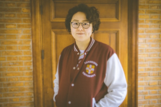 A person with curly hair and glasses is wearing a maroon and white varsity jacket standing in front of a wooden door and brick wall.
