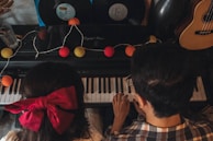Two people are playing a keyboard together, surrounded by colorful string lights and vinyl records. A large red bow is worn by one person whose back is facing the viewer, and a guitar is visible on the side.