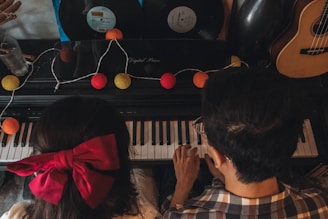 Two people are playing a keyboard together, surrounded by colorful string lights and vinyl records. A large red bow is worn by one person whose back is facing the viewer, and a guitar is visible on the side.