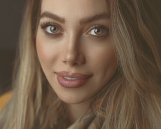 A close-up portrait of a woman with long hair and well-defined facial features. Her eyes are large and expressive, accompanied by full lips and neatly arched eyebrows. The lighting is soft, highlighting her smooth skin and creating a warm atmosphere.