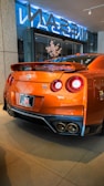 A sleek, orange sports car with a prominent rear spoiler sits in a showroom. The back of the car features dual round tail lights and a visible Nissan GT-R logo. The polished surface of the car reflects the indoor lighting. The showroom has large glass windows and a display with flowers.