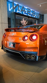 A sleek, orange sports car with a prominent rear spoiler sits in a showroom. The back of the car features dual round tail lights and a visible Nissan GT-R logo. The polished surface of the car reflects the indoor lighting. The showroom has large glass windows and a display with flowers.