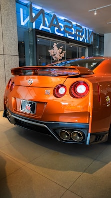 A sleek, orange sports car with a prominent rear spoiler sits in a showroom. The back of the car features dual round tail lights and a visible Nissan GT-R logo. The polished surface of the car reflects the indoor lighting. The showroom has large glass windows and a display with flowers.