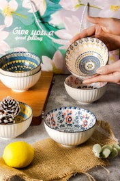 Several ceramic bowls with intricate floral and geometric patterns are arranged on a table. A hand is holding one bowl, showcasing its detailed design. The background includes a decorative paper bag with floral imagery and the words 'Give you a good feeling' written on it. The setting also features a wooden cutting board, a lemon, a pine cone, and a piece of burlap fabric.