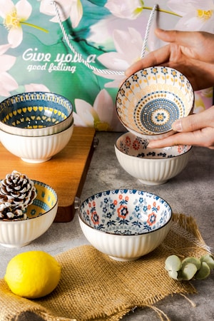 Several ceramic bowls with intricate floral and geometric patterns are arranged on a table. A hand is holding one bowl, showcasing its detailed design. The background includes a decorative paper bag with floral imagery and the words 'Give you a good feeling' written on it. The setting also features a wooden cutting board, a lemon, a pine cone, and a piece of burlap fabric.