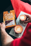 A person holding a package of Oak & Bond Coffee Co., labeled as the Secret Admirer Blend, while two lattes with a heart-shaped latte art and sugar-rimmed glasses sit on a marble slab.