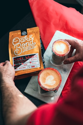 A person holding a package of Oak & Bond Coffee Co., labeled as the Secret Admirer Blend, while two lattes with a heart-shaped latte art and sugar-rimmed glasses sit on a marble slab.