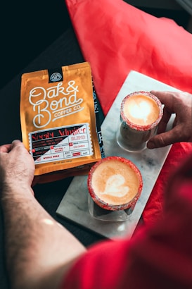 A person holding a package of Oak & Bond Coffee Co., labeled as the Secret Admirer Blend, while two lattes with a heart-shaped latte art and sugar-rimmed glasses sit on a marble slab.