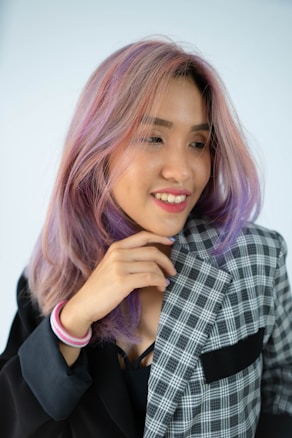 A person with pastel-colored hair, primarily shades of pink and purple, smiles while looking slightly to the side. They are wearing a mixed-patterned blazer with one side in solid black and the other side in a checkered pattern. A pink bracelet adorns their wrist.