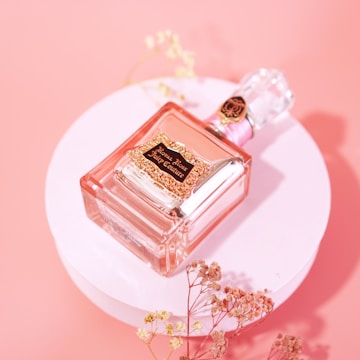 A glass perfume bottle with a decorative golden label lies on a pink circular platform. Delicate dried flowers are arranged around the bottle, complementing the soft pastel background.