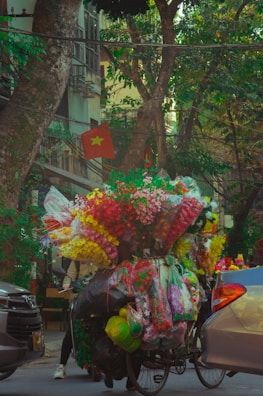 A street scene features a bicycle heavily loaded with vibrant artificial flowers packed in plastic bags. The surrounding environment includes trees, urban architecture, and vehicles. The area appears lively and colorful.