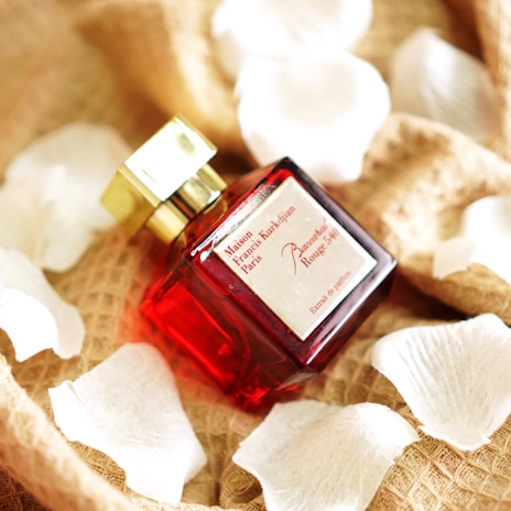 A luxurious red glass perfume bottle with a golden cap is placed on a textured fabric surface. Surrounding the bottle are soft white petals, adding to the sophisticated and elegant presentation.