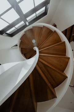 A spiral staircase made of dark wood with a white railing spirals upwards beneath a skylight. The angle creates a dynamic view, emphasizing the curves and geometric design.