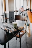 A cozy café setting with a table in the foreground holding a laptop, a green Starbucks drink, a camera, AirPods, and other personal items. A tote bag hangs on the chair. In the background, two people are seated with laptops near large windows, creating a tranquil atmosphere.