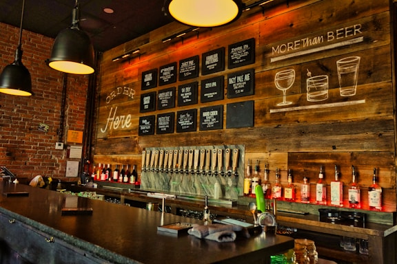 A cozy bar interior with a long counter in front. The wall is decorated with wooden panels and chalkboard-style signs listing various drinks, including beers and cocktails. Several black pendant lights hang from the ceiling, and the shelves behind the bar are lined with rows of liquor bottles. The atmosphere is warm and inviting, enhanced by the soft lighting and rustic decor.