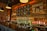 A cozy bar interior with a long counter in front. The wall is decorated with wooden panels and chalkboard-style signs listing various drinks, including beers and cocktails. Several black pendant lights hang from the ceiling, and the shelves behind the bar are lined with rows of liquor bottles. The atmosphere is warm and inviting, enhanced by the soft lighting and rustic decor.