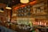 A cozy bar interior with a long counter in front. The wall is decorated with wooden panels and chalkboard-style signs listing various drinks, including beers and cocktails. Several black pendant lights hang from the ceiling, and the shelves behind the bar are lined with rows of liquor bottles. The atmosphere is warm and inviting, enhanced by the soft lighting and rustic decor.
