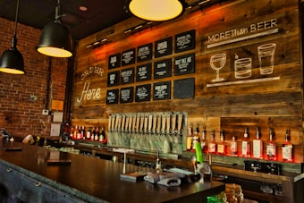 A cozy bar interior with a long counter in front. The wall is decorated with wooden panels and chalkboard-style signs listing various drinks, including beers and cocktails. Several black pendant lights hang from the ceiling, and the shelves behind the bar are lined with rows of liquor bottles. The atmosphere is warm and inviting, enhanced by the soft lighting and rustic decor.