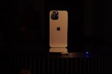 A smartphone with a gold-colored body is placed vertically on a dark surface. The back of the device is visible, featuring a distinctive logo and a multi-lens camera setup at the top left corner. The lighting is low, casting a soft glow on the phone and creating a dramatic contrast with the dark background.