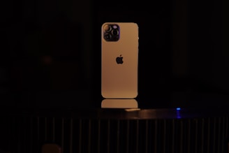 A smartphone with a gold-colored body is placed vertically on a dark surface. The back of the device is visible, featuring a distinctive logo and a multi-lens camera setup at the top left corner. The lighting is low, casting a soft glow on the phone and creating a dramatic contrast with the dark background.