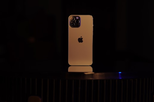 A smartphone with a gold-colored body is placed vertically on a dark surface. The back of the device is visible, featuring a distinctive logo and a multi-lens camera setup at the top left corner. The lighting is low, casting a soft glow on the phone and creating a dramatic contrast with the dark background.