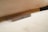 A smartphone lies face down on a light-colored surface, positioned near the edge. The device's metal frame and charging port are visible, with small grill holes likely for speakers and a microphone. The background includes a soft fabric texture, suggesting a couch or bed, contributing to a cozy and relaxed setting.