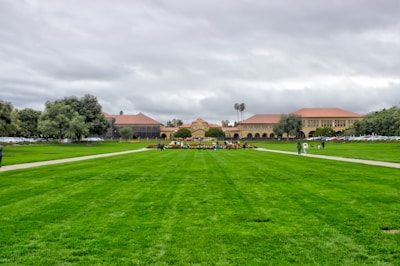 A large open green field is lined by paved walkways, leading towards a complex of historic university buildings with red-tiled roofs. People are casually walking and gathering around the field, with cars parked along the side. The sky is overcast, creating a grayish backdrop against the deep green grass.