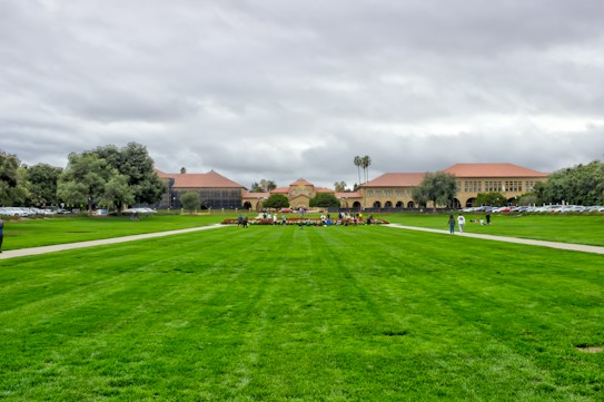 A large open green field is lined by paved walkways, leading towards a complex of historic university buildings with red-tiled roofs. People are casually walking and gathering around the field, with cars parked along the side. The sky is overcast, creating a grayish backdrop against the deep green grass.