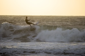 A surfer rides a wave on the ocean, with sunlight casting a warm glow over the scene. The water is choppy, and the surfer is skillfully balancing on the board amidst the crashing waves.
