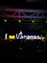 Brightly lit neon sign displaying 'I ♥ Varanasi' with a heart symbol and a flame. The background is dark, highlighting the illuminated letters. Several auto rickshaws are parked in front of the sign.