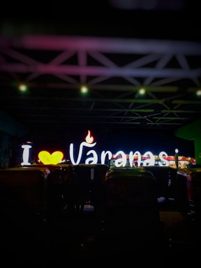 Brightly lit neon sign displaying 'I ♥ Varanasi' with a heart symbol and a flame. The background is dark, highlighting the illuminated letters. Several auto rickshaws are parked in front of the sign.