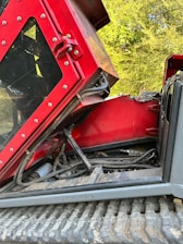 A close-up view of a red mechanical excavator with part of its engine and hydraulic system exposed. The vehicle is resting on a dirt path with trees in the background. Various cables and hoses are visible alongside the machinery components.