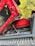 A close-up view of a red mechanical excavator with part of its engine and hydraulic system exposed. The vehicle is resting on a dirt path with trees in the background. Various cables and hoses are visible alongside the machinery components.