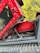 A close-up view of a red mechanical excavator with part of its engine and hydraulic system exposed. The vehicle is resting on a dirt path with trees in the background. Various cables and hoses are visible alongside the machinery components.
