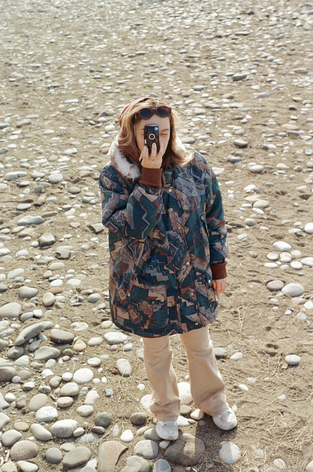 a woman standing on a rocky beach with a camera in her mouth